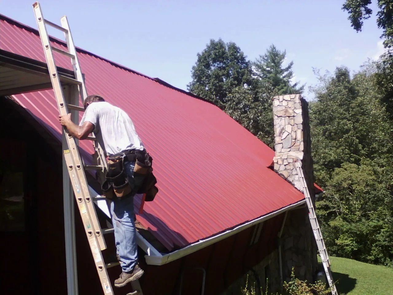 Roofer on a ladder beside a red metal roof and stone chimney — Soffit & Fascia Repair by Roofing Brothers in Hoagland